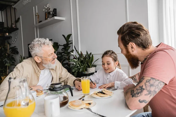 Positive girl writing on notebook near homosexual parents and pancakes at home — Stock Photo