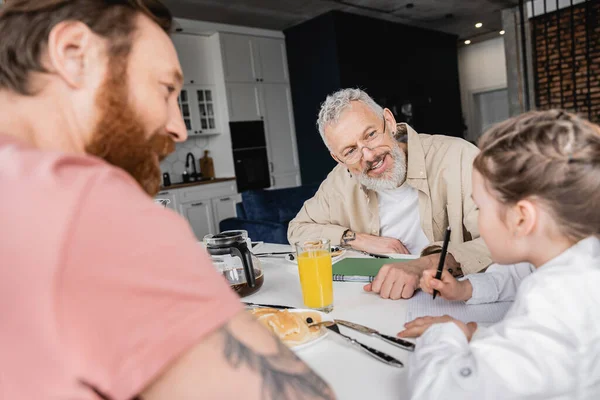 Sorridente gay padri guardando figlia scrittura su notebook vicino colazione a casa — Foto stock