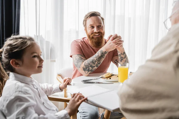 Sonriente gay hombre mirando borrosa pareja cerca preadolescente hija y desayuno en casa - foto de stock