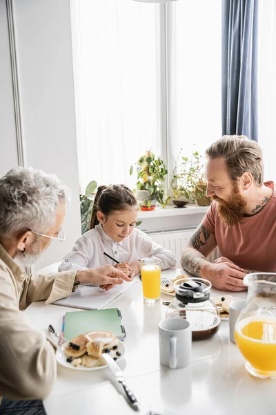 Padres homosexuales mirando a su hija escribiendo en un cuaderno cerca del desayuno y bebidas en casa - foto de stock