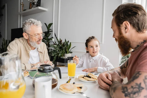 Preteen girl talking to homosexual fathers near notebook and pancakes at home — Stock Photo