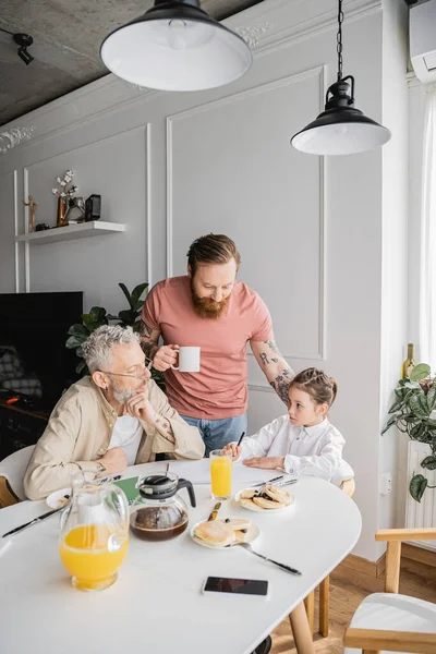 Homosexual parents looking at daughter writing on notebook during breakfast at home — Stock Photo