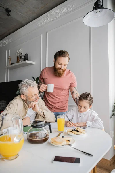 Gay fathers looking at preteen child writing on notebook near breakfast and drinks at home — Stock Photo