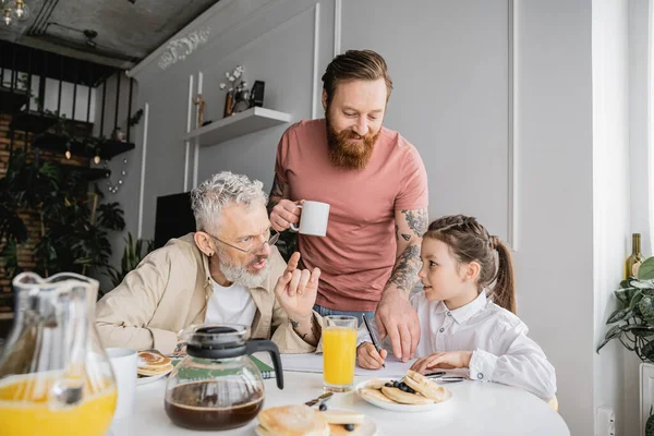 Homosexual parents talking to daughter writing on notebook during breakfast at home — Stock Photo