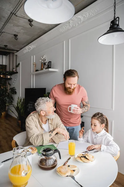 Tattooed gay man holding cup near partner and daughter with notebook during breakfast at home — Stock Photo