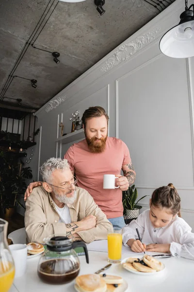 Homosexual parents looking at daughter writing on notebook near pancakes and drinks at home — Stock Photo