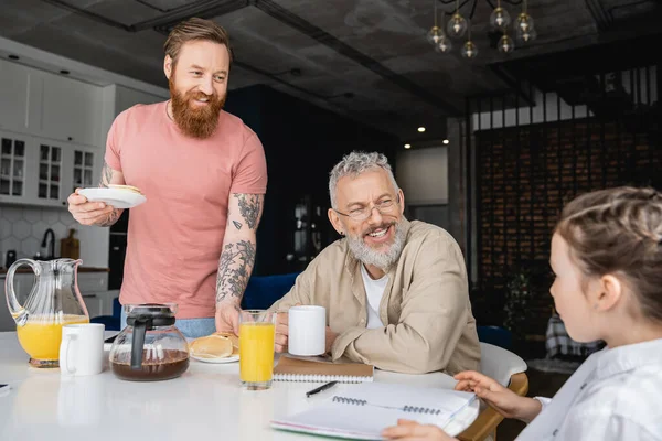 Smiling homosexual parents looking at daughter with notebook during breakfast at home — Stock Photo