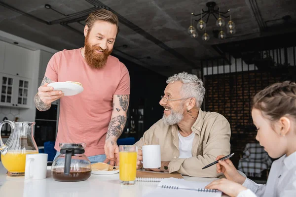 Homosexual man serving pancakes near gay partner and daughter with notebook at home — Stock Photo