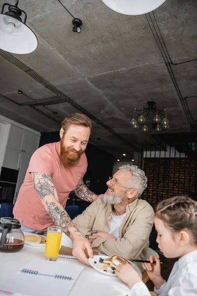 Веселий гей чоловік кладе тарілку з млинцями на стіл біля дочки і партнера вдома — стокове фото
