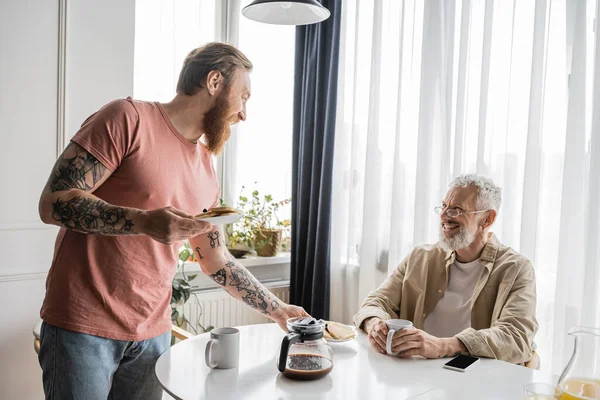 Smiling gay man looking at tattooed partner holding pancakes at home — Stock Photo