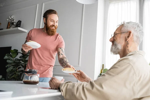 Tätowierter schwuler Mann lächelt, während er seinem Partner zu Hause Pfannkuchen gibt — Stockfoto