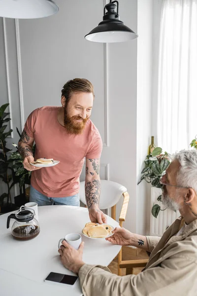 Cheerful tattooed homosexual man giving pancakes to partner at home — Stock Photo