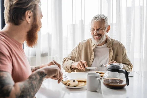 Веселий гей-чоловік розмовляє з розмитим татуйованим партнером під час сніданку вдома — стокове фото
