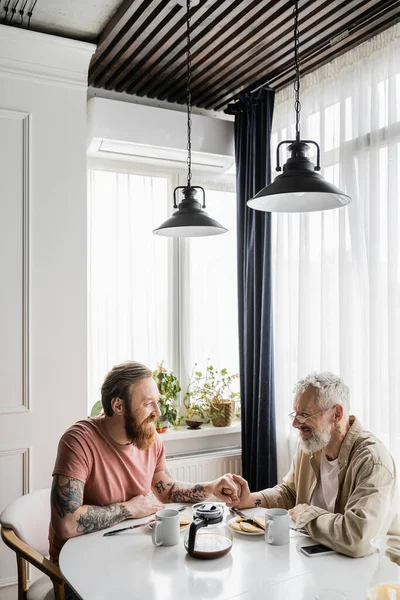 Tätowiertes gleichgeschlechtliches Paar hält Händchen bei Frühstück und Smartphone zu Hause — Stockfoto