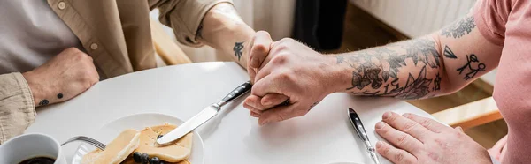 Vista recortada de pareja homosexual tatuada cogida de la mano cerca de café y panqueques en casa, pancarta - foto de stock
