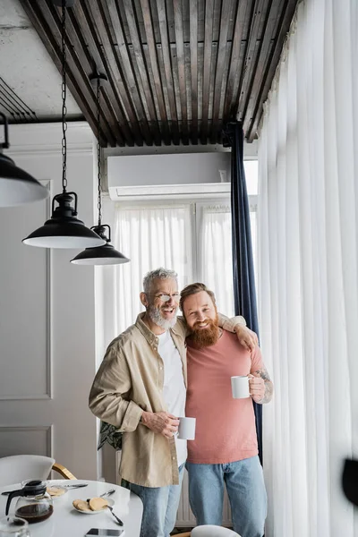 Alegre gay casal abraçando e segurando xícaras de café perto de café da manhã em casa — Fotografia de Stock
