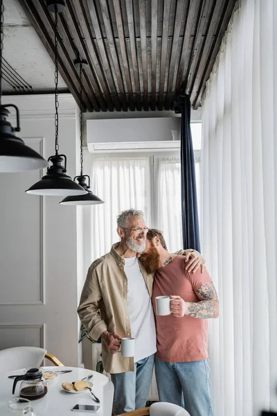Беззаботный гомосексуальный мужчина обнимает татуированного партнера с кофе возле завтрака дома — стоковое фото