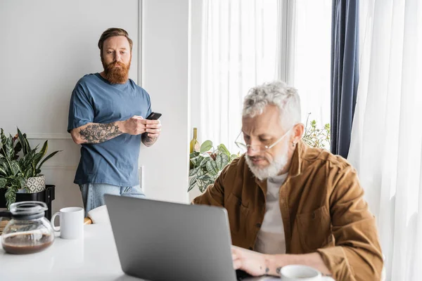 Tattooed gay man using smartphone near partner with laptop and coffee at home — Stock Photo