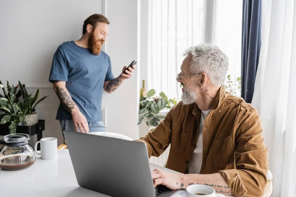 Mature gay man using laptop and looking at blurred partner with cellphone at home — Stock Photo
