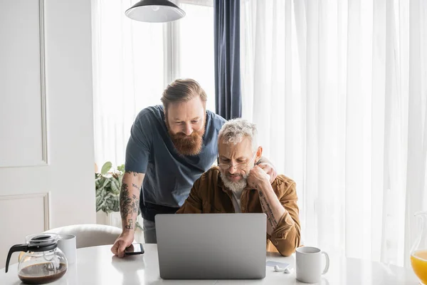 Tattooed gay man hugging mature partner using laptop near coffee at home — Stock Photo