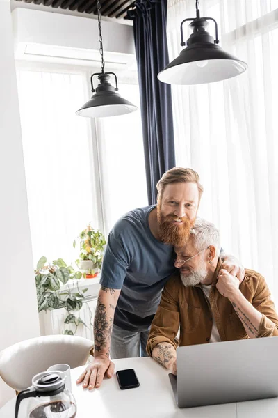 Беззаботный гомосексуальный мужчина обнимает зрелого партнера возле кофе и устройств дома — стоковое фото