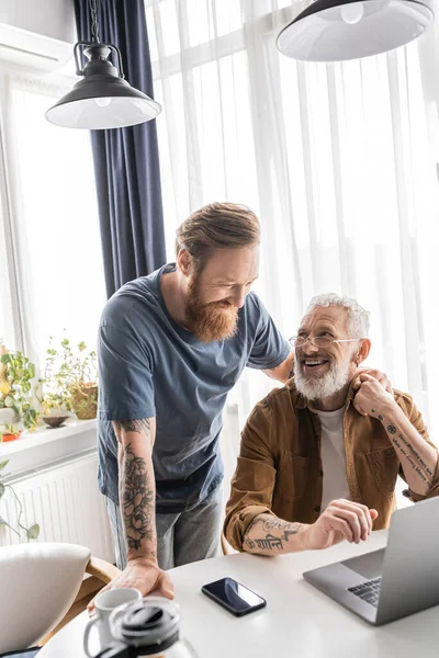 Cheerful gay couple holding hands near gadgets and coffee at home — Stock Photo