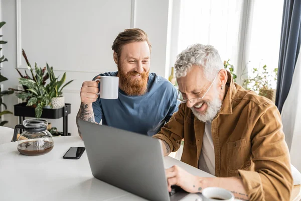 Smiling gay man holding cup of coffee near boyfriend during laptop at home — Stock Photo