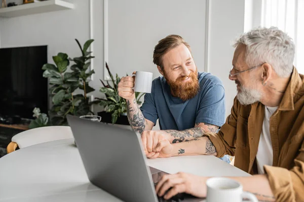 Positive tattooed gay man holding coffee and hand of partner near laptop at home — Stock Photo