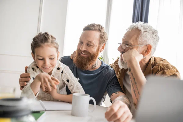 Fröhlich gay eltern looking at preteen daughter near coffee und laptop at home — Stockfoto