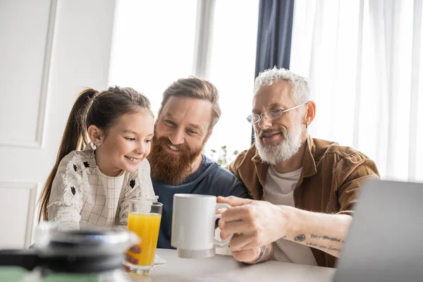 Smiling kid holding orange juice near homosexual parents and laptop at home — Stock Photo