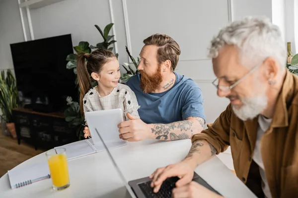Sonriente hombre gay e hija adoptiva usando tableta digital cerca de la pareja con el ordenador portátil en casa - foto de stock