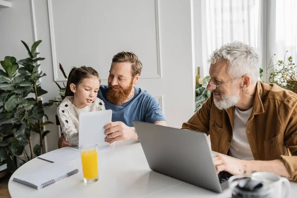 Smiling homosexual man using laptop with daughter near partner with laptop at home — Stock Photo