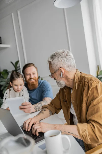 Tattooed gay uomo utilizzando laptop vicino offuscata famiglia con digitale tablet a casa — Foto stock