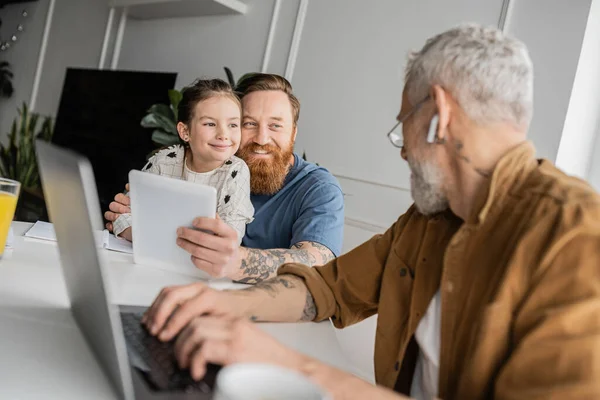 Smiling gay man hugging daughter and holding digital tablet near partner with laptop at home — Stock Photo