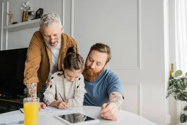 Gay fathers looking at daughter doing homework near digital tablet and orange juice in kitchen — Stock Photo