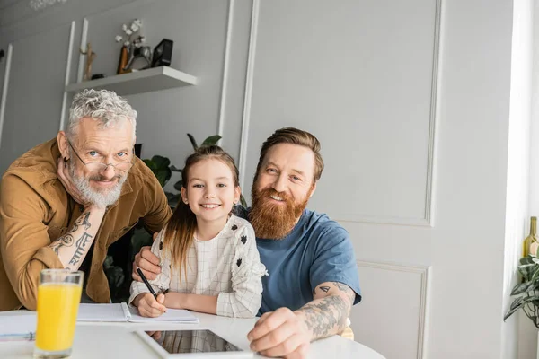 Smiling gay parents with daughter looking at camera while doing homework at home — Stock Photo
