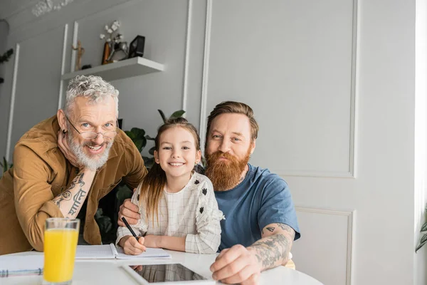 Lächelnde schwule Eltern und ihre Tochter blicken zu Hause auf Kamera in der Nähe von Notebook und digitalem Tablet — Stockfoto