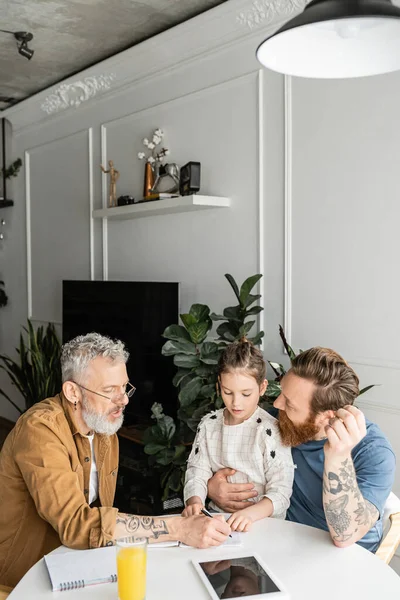 Homosexual parents doing homework with preteen daughter near orange juice and digital tablet at home — Stock Photo