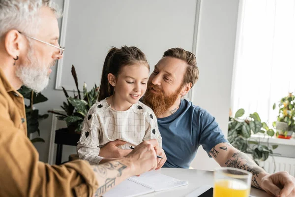 Smiling homosexual father hugging daughter near partner and notebook on table at home — Stock Photo