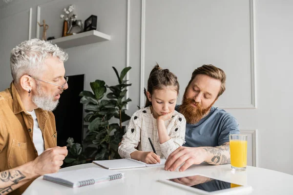 Same sex parents doing homework with daughter writing on notebook at home — Stock Photo