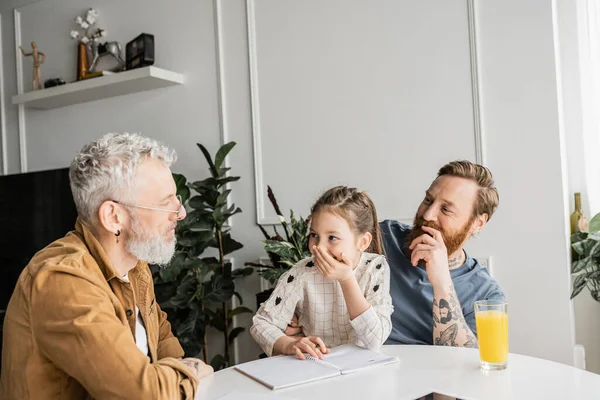 Alegre preadolescente chica haciendo tarea con gay padres cerca naranja jugo en casa - foto de stock