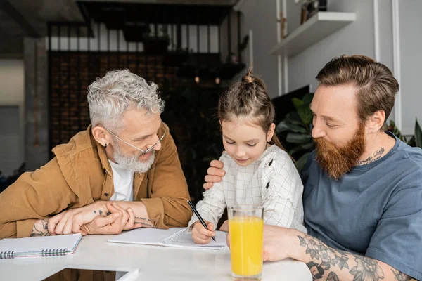 Preteen girl doing homework with same sex parents near notebooks and digital tablet at home — Stock Photo