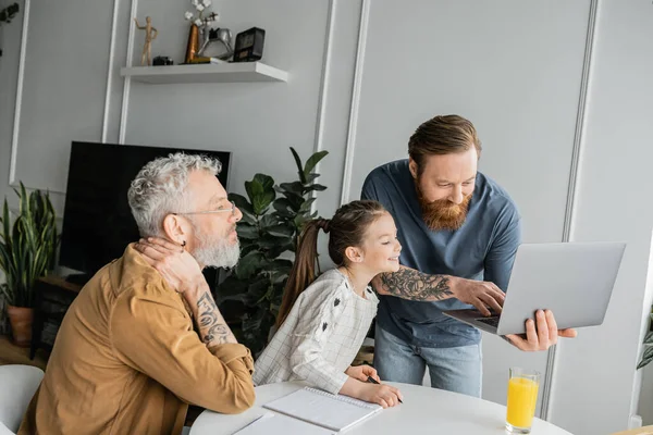 Smiling gay man holding laptop near daughter with notebook and partner at home — Stock Photo
