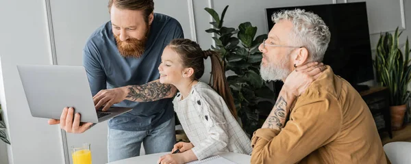 Padres homosexuales con laptop ayudando a su hija sonriente a hacer los deberes en casa, pancarta - foto de stock