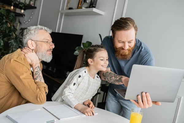 Cheerful kid doing homework near tattooed gay parents with laptop at home — Stock Photo