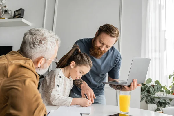 Homosexual man holding laptop while helping daughter with homework near mature partner at home — Stock Photo