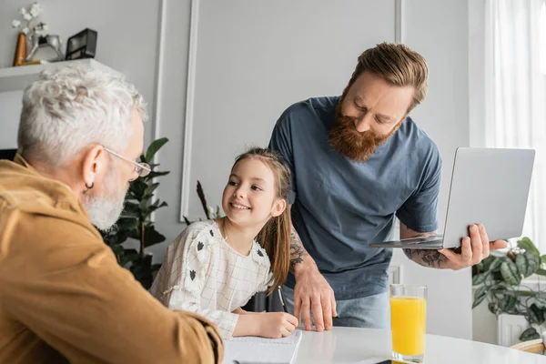 Smiling girl doing homework with same sex parents during online education at home — Stock Photo