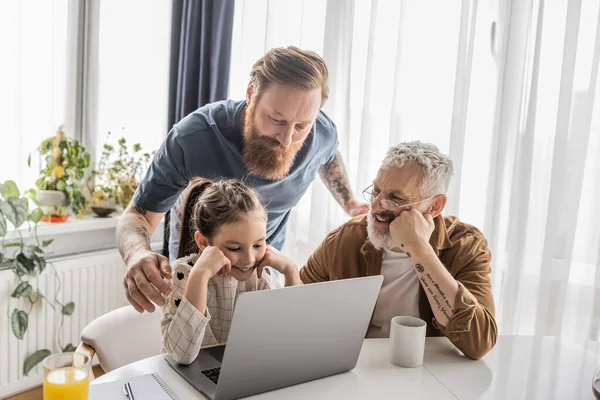 Homosexual fathers using laptop with carefree kid during online education near drinks at home — Stock Photo