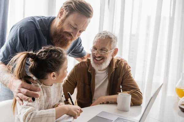 Smiling gay man hugging partner and daughter with notebook near laptop at home — Stock Photo
