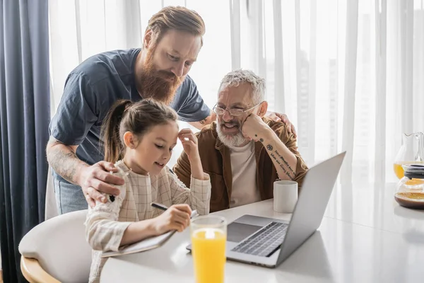 Positive gay men helping daughter with homework near laptop and drinks at home — Stock Photo